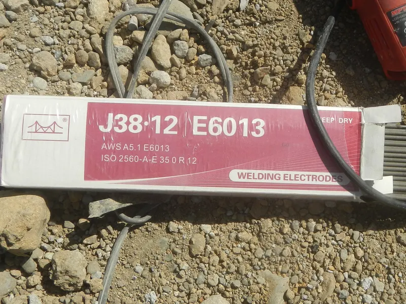 Boîte d'électrodes enrobées E6013 posée sur le sol d'un chantier, à côté d'une pince porte-électrode
