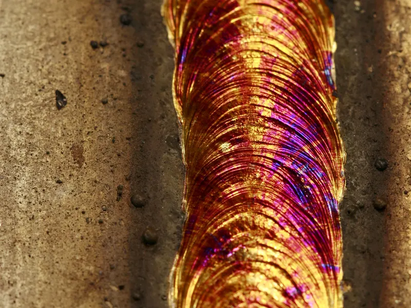 Gros plan d'un cordon de soudure sur acier inoxydable aux reflets dorés, violets et bleus