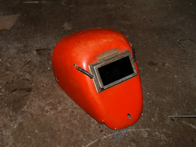 Masque de soudeur à main orange avec sa fenêtre de verre teinté, posé sur un sol d'atelier