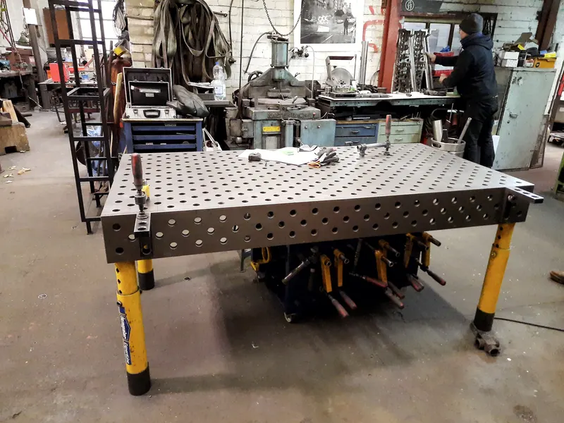 Table de soudage en acier perforée sur pieds jaunes, au milieu d'un atelier de métallerie